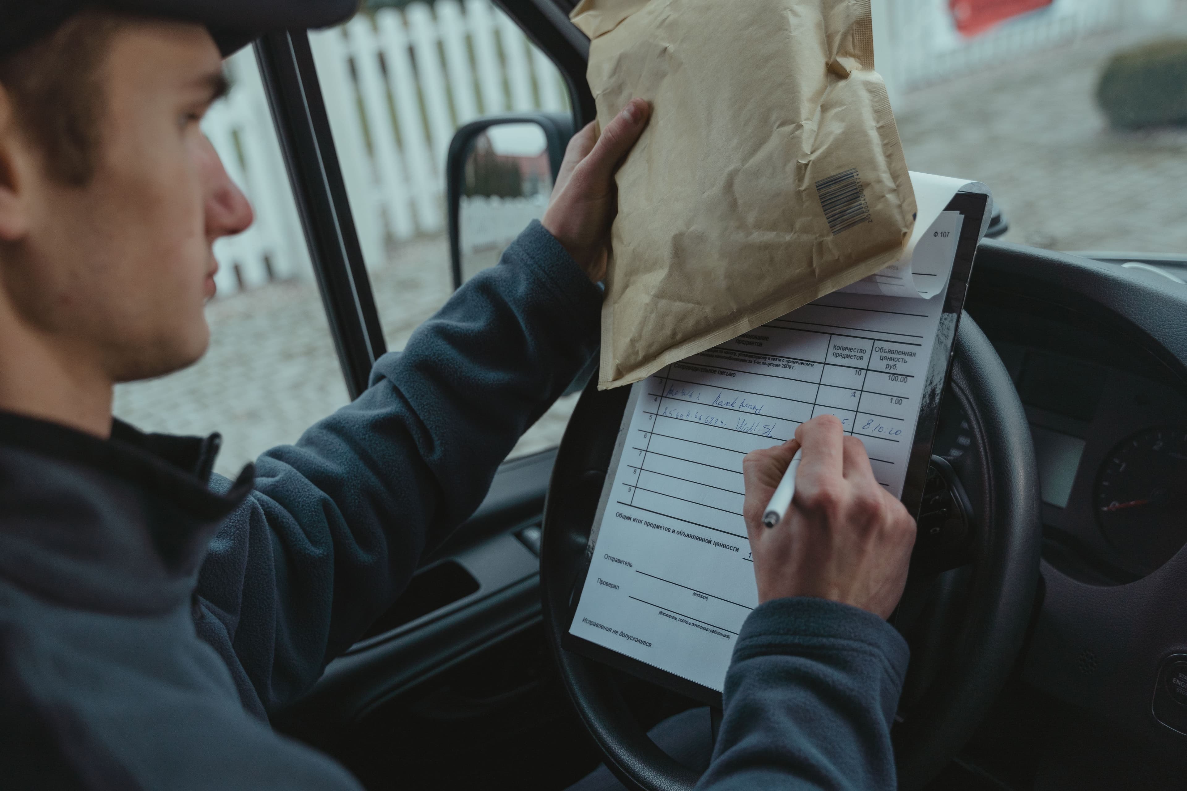A delivery driver parked at the kerb, balancing a parcel on the steering wheel while filling out a paper run sheet by hand